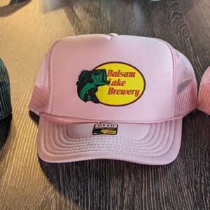 Three trucker hats with the "Bass Pro Shop Balsam Lake Brewery" logo, one green and two pink, displayed on a wooden surface.