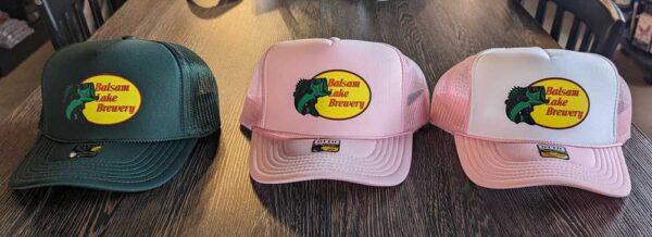 Three trucker hats with the "Bass Pro Shop Balsam Lake Brewery" logo, one green and two pink, displayed on a wooden surface.