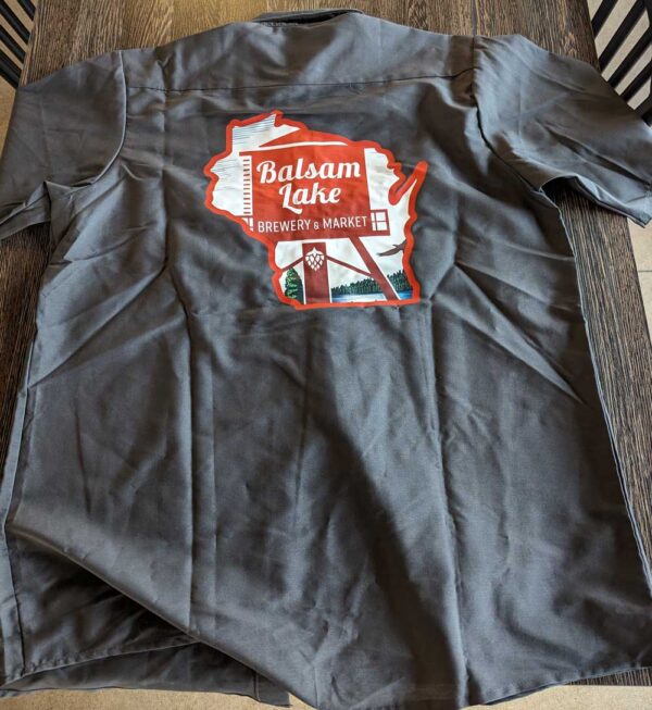 A Brewer's Shirt w/ WI Logo Grey with the balsam lake brewery & market logo printed on it, laid out on a striped surface.