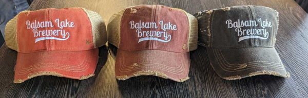 Three Distressed Baseball Caps with "Balsam Lake Brewery" embroidered on the front, displayed on a wooden surface.