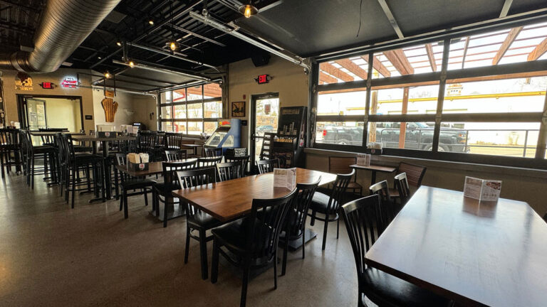 Interior of an empty restaurant with tables and chairs, natural light coming through large windows.