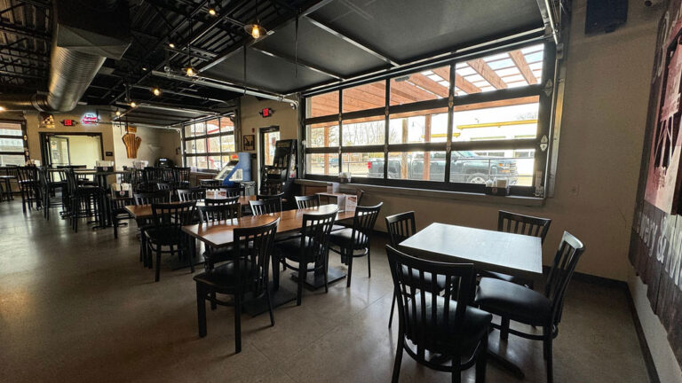 Interior of an empty restaurant with tables and chairs, natural light coming through large windows.