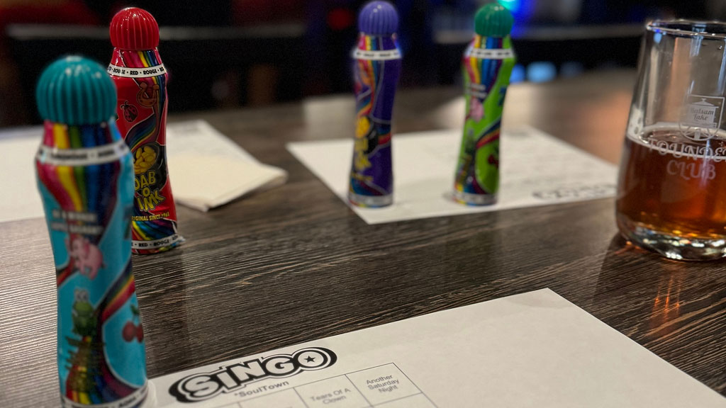 Colorful bingo daubers on a table with a 'singo' game card and a half-empty glass of beer in the background.