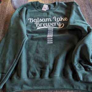 Crewneck Sweatshirt Green with "balsam lake brewery" printed on the front, displayed on a wooden table.