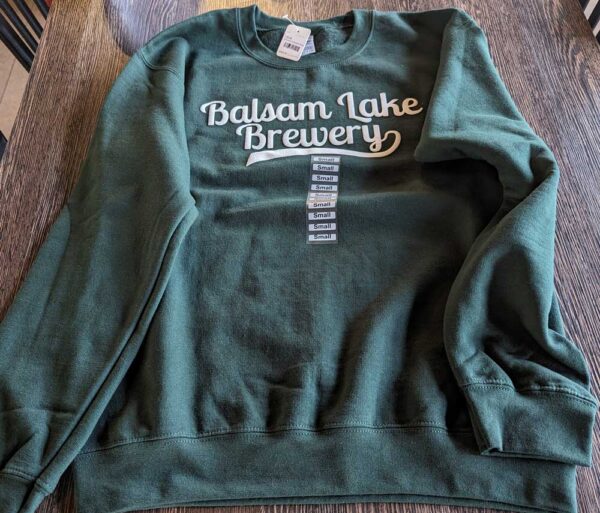 Crewneck Sweatshirt Green with "balsam lake brewery" printed on the front, displayed on a wooden table.