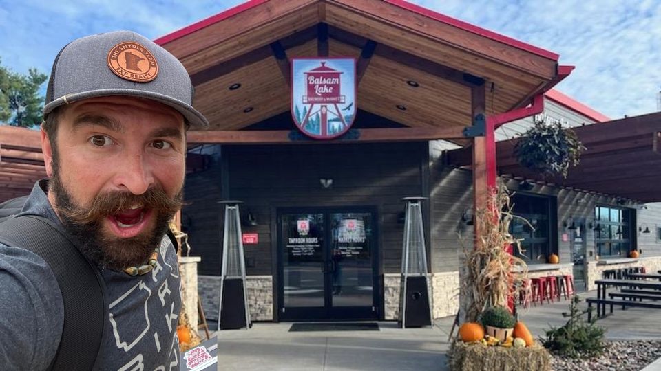 A man with a beard and mustache wearing a cap and holding a drink stands in front of a restaurant named "Balsam Lake Brewing Company.