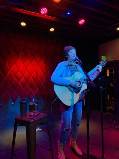 A person wearing a beanie plays an acoustic guitar and sings into a microphone on a dimly lit stage.