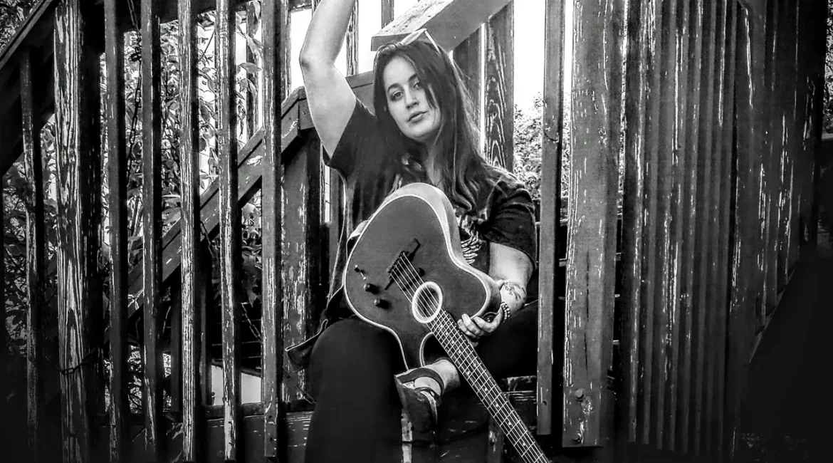 Person sitting with a guitar on a wooden staircase outdoors in black and white.