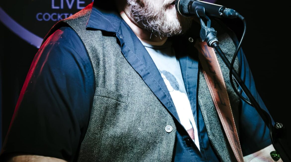 A bearded man wearing a hat, sunglasses, and a vest sings into a microphone while playing an acoustic guitar on stage.