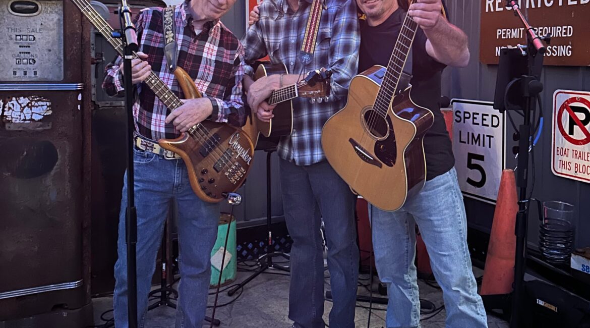 Three men stand together holding guitars and smiling on an outdoor stage, surrounded by music equipment and street signs.