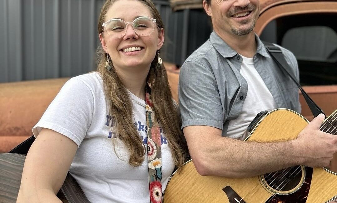 Two people stand outdoors holding guitars and smiling, with an old, rusted vehicle in the background.