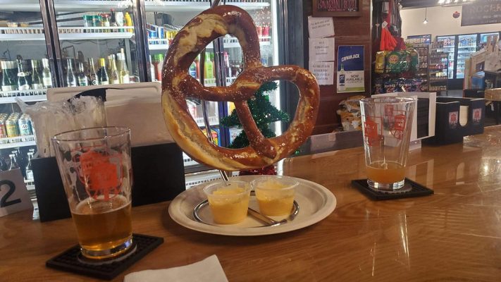 A large pretzel on a stand accompanied by two small cups of sauce and two half-consumed beers on a bar counter.